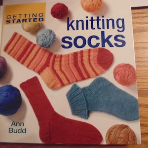 Puede incluir: Un libro titulado "Getting Started Knitting Socks" de Ann Budd. La portada presenta calcetines de punto de colores y ovillos de lana.