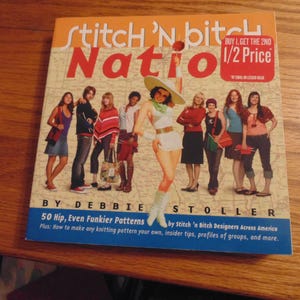 Puede incluir: Portada del libro "Stitch 'N Bitch Nation" con un grupo de personas. La portada es naranja y azul con el texto "Buy 1, Get the 2nd 1/2 Price". El libro es de Debbie Stoller y contiene patrones de tejido.