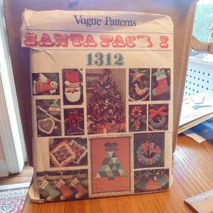 May include: A white paper envelope for a Vogue Patterns sewing pattern labelled 1312 Santa Pac 3. The front features multiple colour photographs of finished holiday-themed fabric crafts, including stockings, ornaments, a tree skirt, and a wall hanging.