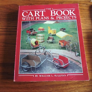 Puede incluir: El libro « The Cart Book with Plans & Projects » de William L. Sullivan. La portada roja presenta el título en blanco y amarillo. La imagen muestra varios diseños de carros, incluyendo carros de bicicleta y carros cubiertos, sobre un césped verde.