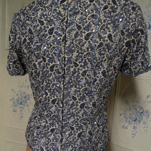 May include: A short-sleeved, beaded top with a paisley pattern in shades of blue and gold. The top has a fitted silhouette and a scoop neckline.