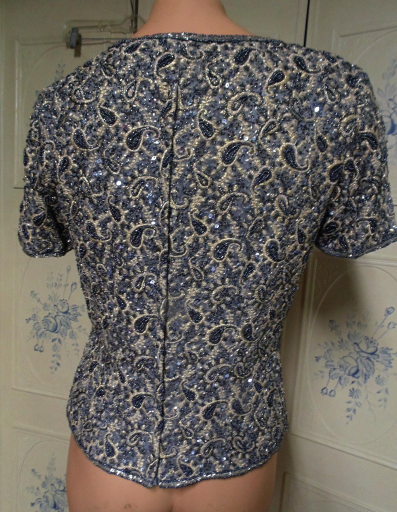 May include: A short-sleeved, beaded top with a paisley pattern in shades of blue and gold. The top has a fitted silhouette and a scoop neckline.