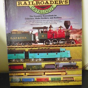 May include: The Model Railroader's Catalogue book cover features a colorful illustration of model trains, including a red and green steam engine, a blue diesel engine, and several freight cars. The text on the cover reads "The Model Railroader's Catalogue" and "The Complete Sourcebook for Collectors, Model Builders, and Railfans".