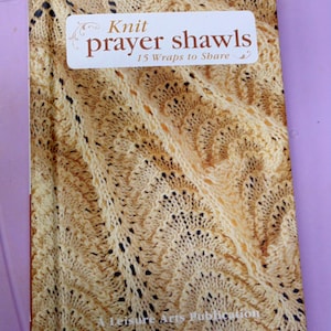 May include: A book titled "Knit prayer shawls" with a beige knitted pattern on the cover. The book features a white label with the title and the phrase "15 Wraps to Share". The cover has a textured, detailed design.