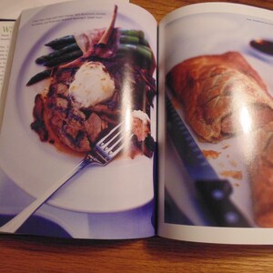 May include: A cookbook spread featuring two plated dishes. One plate holds a grilled meat dish with asparagus and a dollop of white topping. The other shows a golden-brown pastry with a knife.