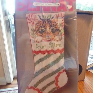 May include: A Bernat Stamped Cross-Stitch kit for a Christmas stocking featuring a brown tabby kitten with a red and white candy cane, green holly leaves, and the name Sue Ellen printed in green script on a white background with green stripes.