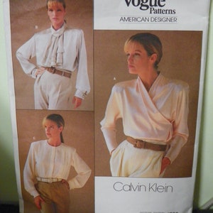May include: A vintage Vogue Patterns sewing pattern for a blouse by Calvin Klein. The pattern is for a white blouse with a wrap style and a ruffled collar. The pattern number is 1792.