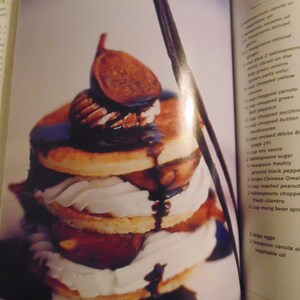 May include: A layered dessert featuring pancakes, whipped cream, figs, and pecans, drizzled with chocolate sauce. The dessert is presented on a white plate, with a dark brown vanilla bean visible.