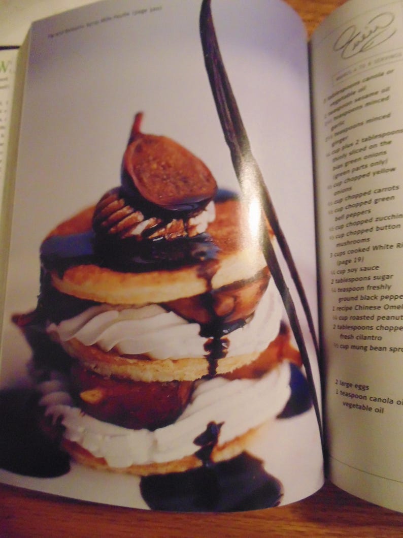 May include: A layered dessert featuring pancakes, whipped cream, figs, and pecans, drizzled with chocolate sauce. The dessert is presented on a white plate, with a dark brown vanilla bean visible.