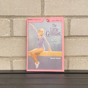 May include: A paperback book titled The Little Gymnast by Sheila Haigh with a pink border. The cover features an illustration of a gymnast in a light purple leotard sitting on a balance beam. The text Apple Paperbacks appears at the top.