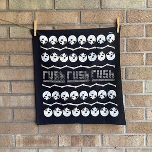 May include: Black bandana with a repeating pattern of white skulls and bones. The text "RUSH RUSH RUSH" is printed in white across the center. The bandana is hanging on a clothesline against a brick wall.