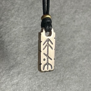 Peut inclure: Un collier avec un pendentif rectangulaire et un cordon noir. Le pendentif est de couleur métallique claire avec un motif gravé d'une ligne verticale avec des flèches et un point. Le cordon est noué en haut avec un anneau doré.