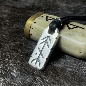 May include: A silver rectangular pendant with etched symbols hangs from a black cord. The pendant features a series of arrows and lines. The background includes a bone-coloured object with brass accents and a dark, textured surface.