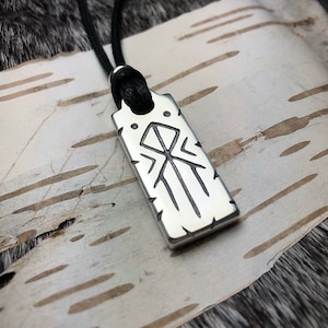 Puede incluir: Un collar con un colgante de plata y un cordón negro. El colgante es rectangular y presenta un símbolo de runa nórdica.
