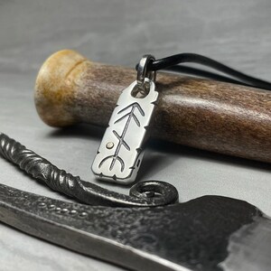 Puede incluir: Collar con colgante de plata y cordón de cuero negro. El colgante es un amuleto rectangular de plata con un diseño de árbol estilizado y un pequeño punto dorado en el centro.