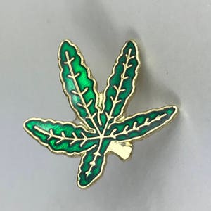 May include: A gold-colored pin with a green enamel marijuana leaf design.