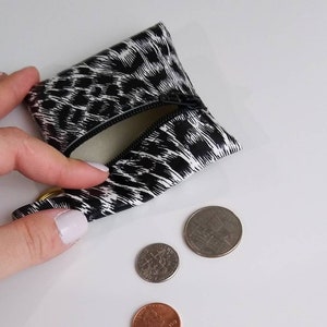 May include: A small, black and white leopard print coin purse with a zipper is open, revealing a light interior. Three coins are next to the purse: a penny, a dime, and a quarter. The purse has a gold-colored ring attached.