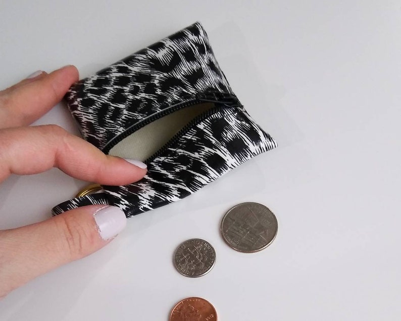 May include: A small, black and white leopard print coin purse with a zipper is open, revealing a light interior. Three coins are next to the purse: a penny, a dime, and a quarter. The purse has a gold-colored ring attached.