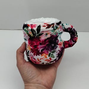 Może przedstawiać: Biały ceramiczny kubek z kwiatowym wzorem w odcieniach r&oacute;żowego, fioletowego, pomarańczowego i zielonego. Kubek jest wypełniony białym wł&oacute;knem.