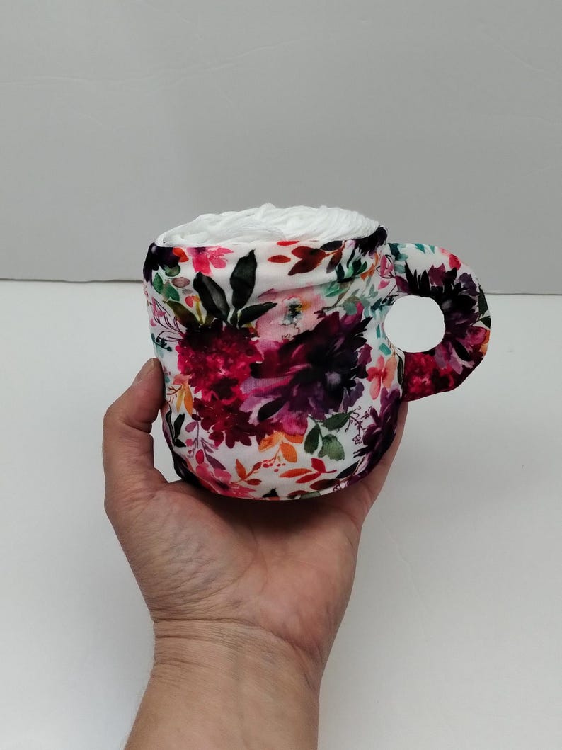 Może przedstawiać: Biały ceramiczny kubek z kwiatowym wzorem w odcieniach r&oacute;żowego, fioletowego, pomarańczowego i zielonego. Kubek jest wypełniony białym wł&oacute;knem.