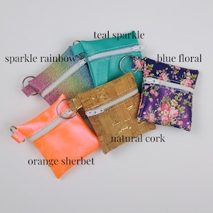 May include: A collection of small, zippered pouches in various colors and patterns. The pouches include "sparkle rainbow", "teal sparkle", "blue floral", "natural cork", and "orange sherbet". Each pouch has a silver zipper and a metal ring.