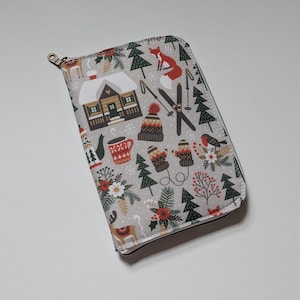 May include: A rectangular zipped case featuring a light grey background with a repeating winter-themed pattern. The design includes illustrations of a snow-covered cabin, a red fox, skis, a knit hat, mittens, a mug, pine trees, and a small bird.