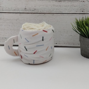 Może przedstawiać: Biały ceramiczny kubek z kolorowym wzorem konfetti, wypełniony kulką białej wł&oacute;czki.