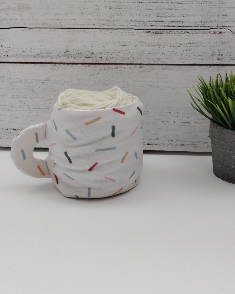 Może przedstawiać: Biały ceramiczny kubek z kolorowym wzorem konfetti, wypełniony kulką białej wł&oacute;czki.