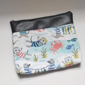 May include: A rectangular pouch with a black leather top and a colorful fabric body. The fabric features a playful design of otters, fish, and a crab in a sea-themed setting. The otters are wearing striped shirts.