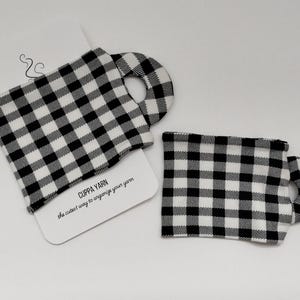 Może przedstawiać: Dwa czarne i białe ginghamowe pojemniki na wł&oacute;czkę. Pojemniki mają kształt kubk&oacute;w z uchwytem. Tekst "CUPPA YARN the cutest way to organize your yarn" jest nadrukowany na białej kartce za pojemnikami.