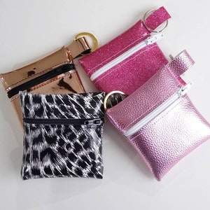 May include: Four small, square coin purses in various colors and textures. One is metallic rose gold, another is hot pink glitter, a third is a textured light pink, and the last has a black and white animal print. Each has a zipper and a key ring.