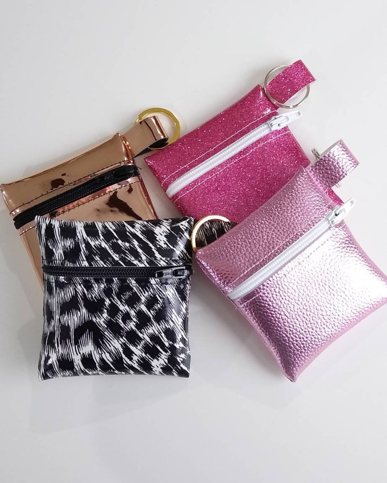 May include: Four small, square coin purses in various colors and textures. One is metallic rose gold, another is hot pink glitter, a third is a textured light pink, and the last has a black and white animal print. Each has a zipper and a key ring.