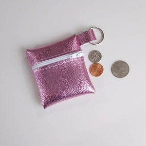 May include: A small, pink coin purse with a textured finish and a white zipper. The purse has a loop with a silver key ring attached. Three coins are next to the purse: a dime, a penny, and a quarter.