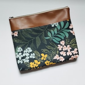 May include: A rectangular pouch with a brown leather top and a floral patterned bottom. The floral pattern features white, yellow, and pink flowers with green leaves on a dark background. A zipper runs along the top.