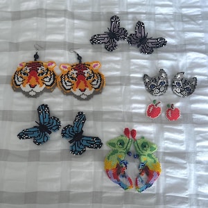 May include: A selection of beaded earrings featuring diverse designs. The collection includes tiger faces, butterflies, cat faces, apples, and chameleon-like creatures. The earrings are colourful and detailed.