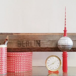 May include: A red and white patterned tin can with paint brushes, a red and white patterned tin can, a wooden sign with the word "BERLIN" on it, a silver and red model of the Berlin TV Tower, and a gold clock.