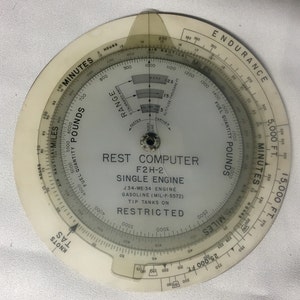 May include: A vintage circular flight computer with a clear plastic overlay. The device features multiple scales for calculating flight data, including nautical miles, fuel quantity in pounds, and altitude. Text on the computer reads "REST COMPUTER F2H-2 SINGLE ENGINE".