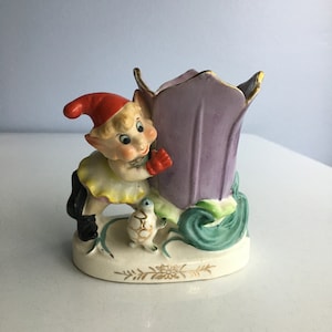May include: A ceramic figurine of a whimsical elf holding a purple flower pot. The elf is wearing a red hat, yellow dress, and black boots. A small turtle is sitting next to the elf.
