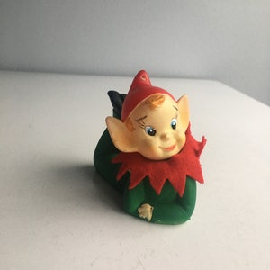 May include: A green and red Christmas elf figurine with a smiling face and a red hat. The elf is lying on its stomach with its arms crossed.
