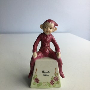 May include: A ceramic figurine of a red-clad elf sitting on a green and white base. The base has the words "Rochester Minn." written on it.
