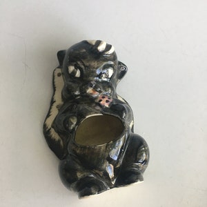 May include: A black and white ceramic skunk planter with a small hole in the top for planting. The skunk is holding a red and white heart in its paws.