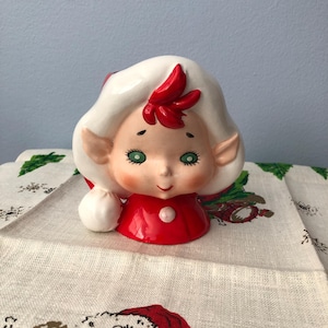 May include: A ceramic Christmas elf figurine with a white hat, red and white outfit, and green eyes. The elf is smiling and has a rosy cheek.