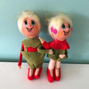 May include: Two vintage felt Christmas ornaments depicting a boy and girl with blonde hair, wearing green pants and red shirts. They have painted faces with rosy cheeks and blue eyes.