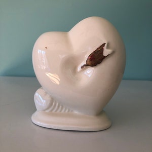 May include: A white ceramic heart-shaped vase with a small base. The vase has a smooth, glossy finish and a decorative, leaf-like accent with brown and red tones. The vase is a decorative item.