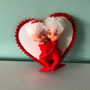 May include: Two red felt dolls with white fuzzy hair are hugging inside a red heart-shaped frame with white trim.