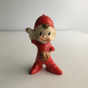 May include: A small ceramic figurine of an elf wearing a red baseball uniform and holding a baseball glove. The elf has a red hat, brown hair, and a smiling face.