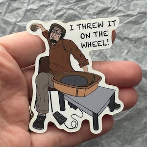 May include: A sticker featuring a cartoon illustration of a person throwing a ball of clay onto a pottery wheel. The person is wearing a brown shirt and cap, with the text "I THREW IT ON THE WHEEL!" in a speech bubble.