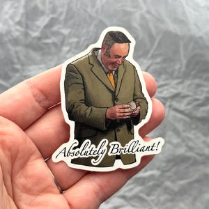 May include: A sticker featuring a cartoon illustration of a man in a green blazer and yellow tie, holding a small object. The text "Absolutely Brilliant!" is written in cursive below the image.
