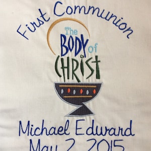 May include: White fabric with blue embroidery that reads "First Communion" and "The Body of Christ" with a stylized chalice. Below the chalice is the name "Michael Edward" and the date "May 2, 2015".