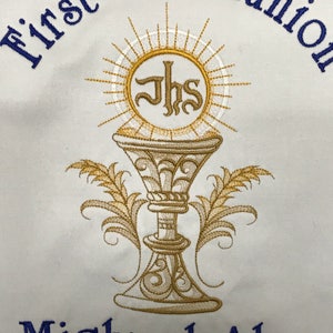 May include: White fabric embroidered with "First Communion" in blue script. A gold chalice with the letters "JHS" is centered, surrounded by a sunburst design. The name "Michael Alan" and date "May 5, 2018" are embroidered below.